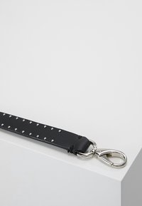 Bracelet en cuir noir avec petits clous argentés et un fermoir mousqueton en métal argenté, reposant sur un coin de surface blanche.