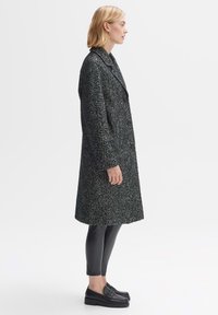 Double-breasted coat in dark textured fabric, featuring a notched collar and side pockets. Paired with shiny black leggings and platform shoes.