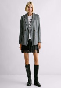 Donna con bob biondo che indossa un blazer grigio doppiopetto, una t-shirt bianca con stampa, una gonna nera con frange e stivali neri al ginocchio.