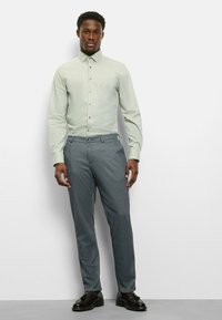 Hellgrünes langärmliges Hemd mit Knopfleiste, kombiniert mit grauer Hose und schwarzen Schuhen. Glatte Textur, schlichte Linienführung und maßgeschneiderte Passform.