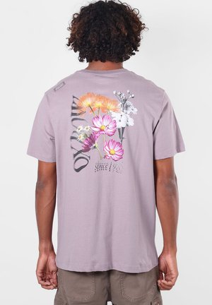 Personne aux cheveux bouclés portant un t-shirt violet clair avec des fleurs colorées et le texte "OXYDITY since 1985" dans le dos.