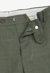 Pantalones verdes texturizados con cremallera abierta, cierre de botón y broche metálico en la cintura, sobre fondo blanco.