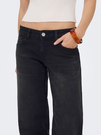 Femme portant un jean noir à jambes larges et un top blanc côtelé court avec un grand bracelet en ambre à son poignet droit.