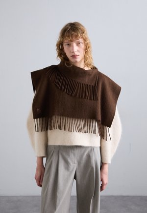 Poncho marrón con flecos y cuello alto sobre un suéter crema, combinado con pantalones de corte claro en gris. Texturas suaves y diseño en capas.