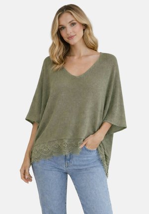 Femme aux cheveux blonds portant un haut ample vert olive à col en V avec bordure en dentelle et un jean bleu clair, debout avec une main dans la poche.