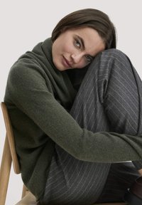 Grüner Wollpullover mit einem drapierten Ausschnitt, kombiniert mit grauen gestreiften Hosen. Model sitzt auf einem Holzstuhl vor einem neutralen Hintergrund.