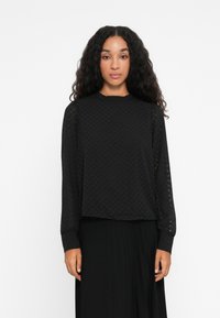 Black long-sleeve top with a textured herringbone pattern, round neckline, and fitted cuffs. Paired with a black pleated skirt.
