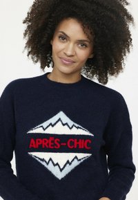Marineblauer Pullover aus weichem Material mit einem Logo, das stilisierte Berge und "APRÈS-CHIC" in Rot und Weiß zeigt. Rundhalsausschnitt, entspannte Passform.