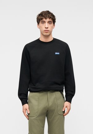 Ung mann med krøllete hår har på seg en svart genser med lange ermer med en liten blå logo og olivengrønne bukser mot en ensfarget bakgrunn.