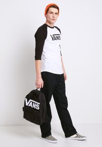 Sort og hvid Vans baseball t-shirt med lange ærmer, sorte bukser, sort rygsæk og ternede slip-on sko, samt en orange hue.