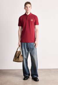 Κόκκινο πόλο μπλουζάκι με κεντητό έμβλημα, σε συνδυασμό με άνετο μπλε τζιν και καφέ δερμάτινη τσάντα. Μαύρα παπούτσια ολοκληρώνουν την εμφάνιση.