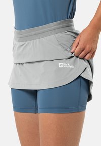 Grijs rokje met een geplooid tailleband en een zijzak, over blauwe sportieve shorts. Jack Wolfskin-logo bedrukt op de zoom.