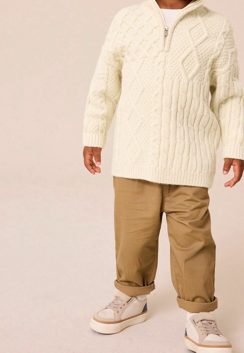 Enfant portant un pull crème à torsades, un pantalon beige retroussé, des chaussettes blanches et des baskets beiges avec des touches bleues, debout sur un sol clair.