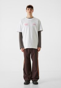 Biela bavlnená tričko s ružovým grafickým textom, vrstvené na dlhom hnedom rebrovanom tričku. Tmavohnedé tepláky s oranžovými bočnými pruhmi.