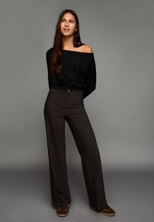 Black off-shoulder top paired with high-waisted, pinstriped, wide-leg trousers and brown suede sneakers. Neutral backdrop.