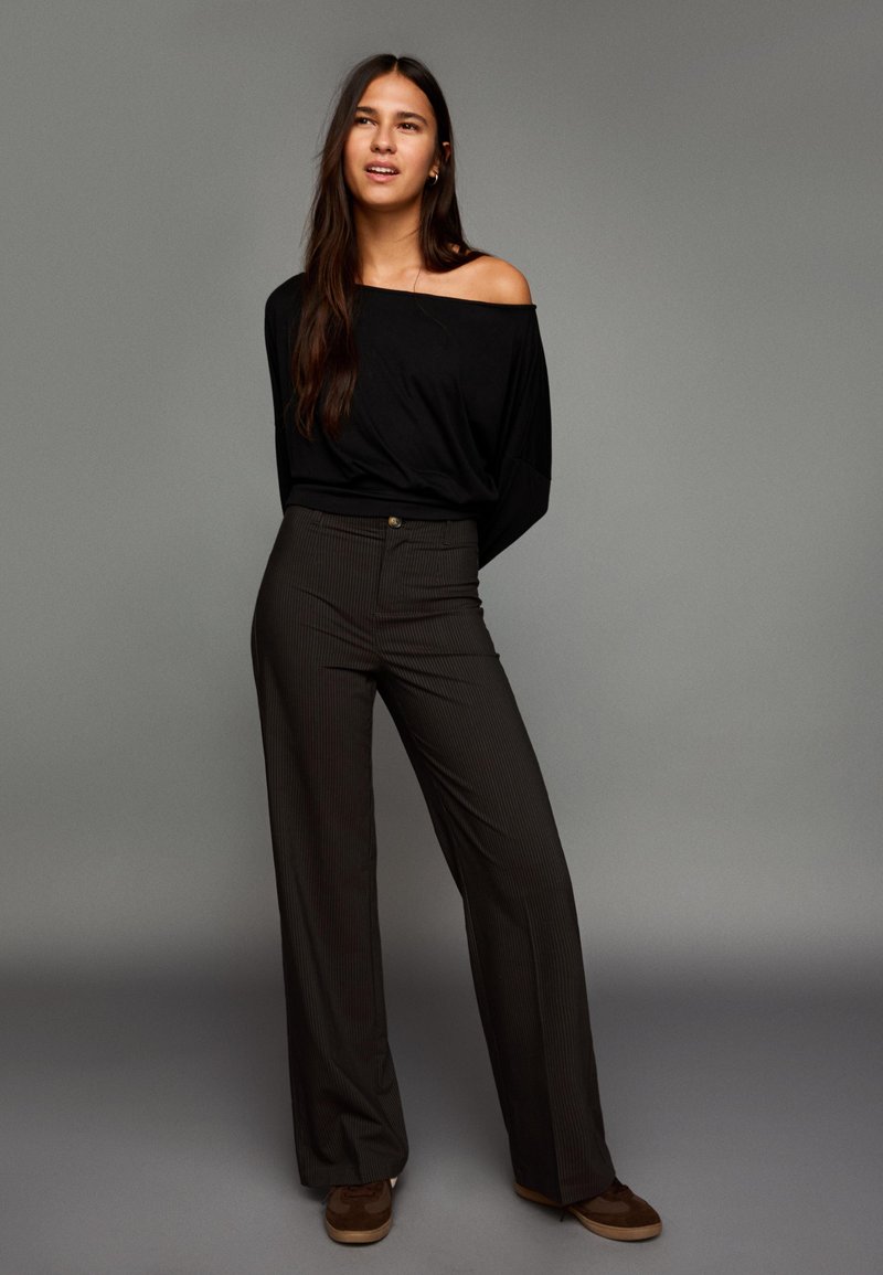 Črna majica brez ramen v kombinaciji z visokim pasom, hlačami širokih nog z ozkimi črtami in rjavimi semišnimi supergami. Nevtralen ozadje.