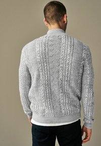 Szary sweter z dzianiny z teksturowanymi wzorami w warkocze, z ribbowym dekoltem i mankietami oraz luźnym krojem, noszony na białej koszuli.