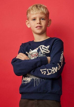 Blonde jongen met gekruiste armen in een marineblauwe graphic sweatshirt die zelfverzekerd tegen een effen rode achtergrond staat.