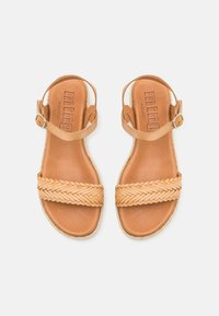 Sandales en cuir tan avec une sangle tressée et une boucle de cheville. La texture lisse et la couleur naturelle créent un design minimaliste.