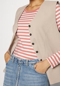 Beige Weste mit strukturiertem Design und schwarzen Knöpfen, getragen über einem rot-weißen gestreiften Hemd, kombiniert mit einem blauen Jeansrock.