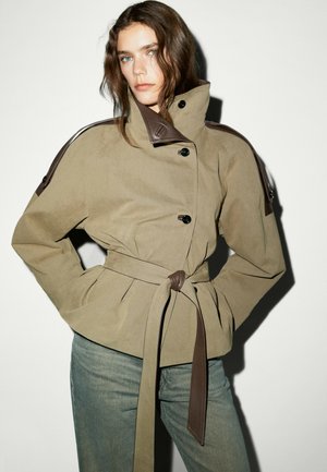 Donna che indossa una giacca beige con cintura, dettagli marrone scuro e collo alto, abbinata a jeans blu, in piedi su sfondo bianco.