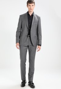 Grå dress med skreddersydd passform, to-knappers jakke, notched lapels og flate bukser. Båret med en svart skjorte med knapper og svarte sko.