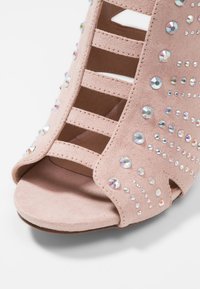 Sandalia abierta de color rosa empolvado que presenta un material de ante suave, múltiples cortes horizontales y está adornada con diferentes tamaños de cristales de vidrio transparentes.