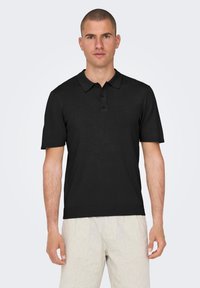Black short-sleeve polo shirt with a collared neck and three-button placket, paired with striped light-colored shorts.