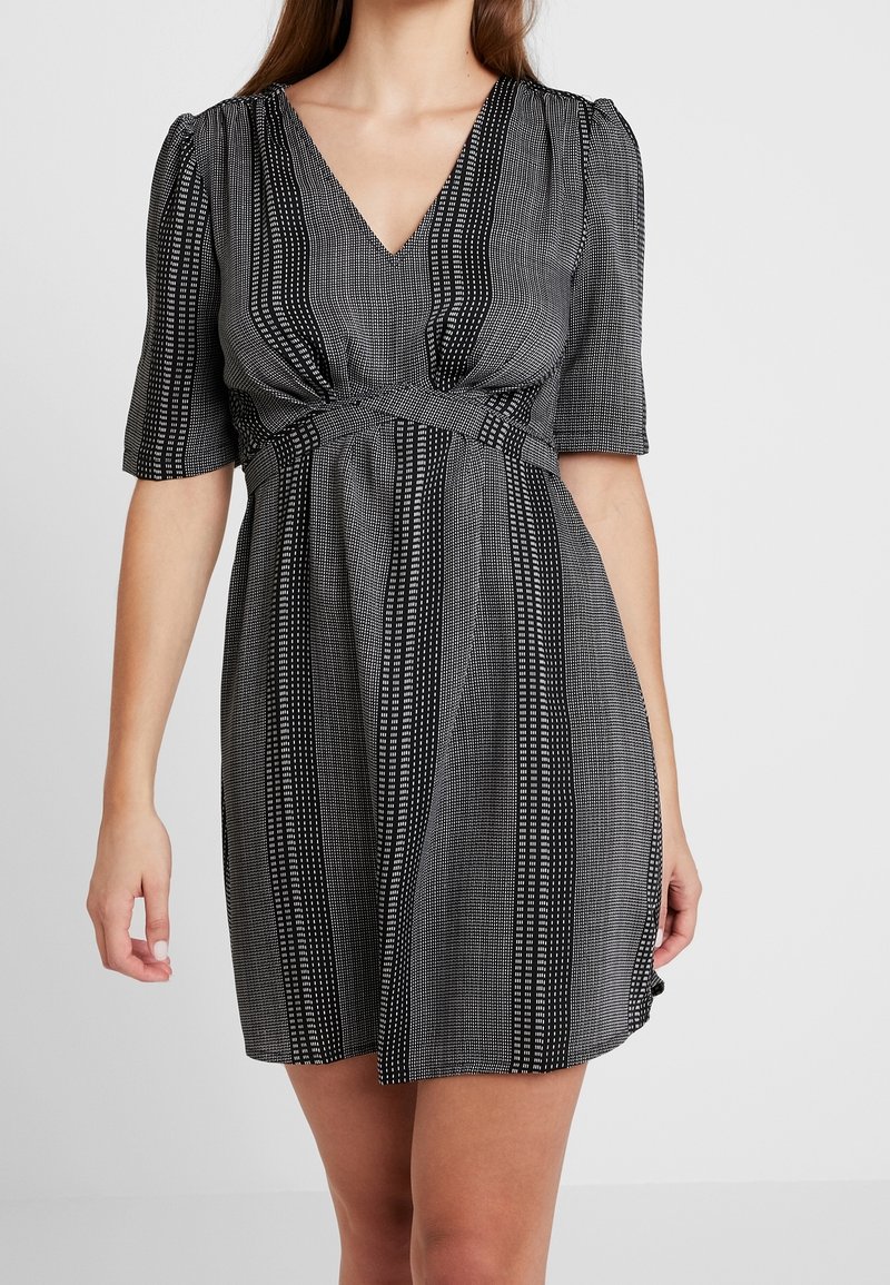 Femme portant une robe courte à motif noir et blanc avec un col en V et des manches aux coudes sur un fond uni.