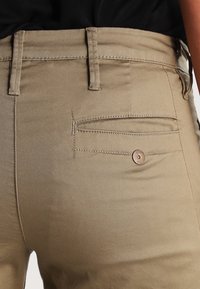 Tan tygbyxor med fram- och bakfickor, utrustade med en knappdetalj på bakfickan och synlig sömnad längs sömmarna.