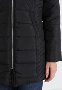 Giacca trapuntata nera con zip frontale, tasca laterale e maniche lunghe indossata sopra jeans blu con mano destra visibile.