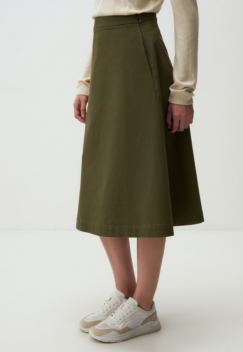 Olijfgroene A-lijn rok met een gladde textuur, hoge taille en naden, gecombineerd met witte en beige sneakers.