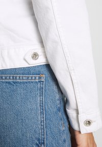 Manche de veste en denim blanc avec boutons-pression métalliques associée à une poche de jean bleu et des détails de couture.