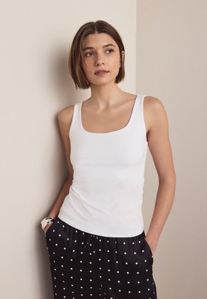 Jauna sieviete ar īsiem brūniem matiem, valkājot baltu tanktopu un melnas bikses ar punktiņiem, stāv ar rokām kabatās un skatās uz sāniem.