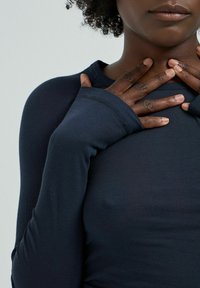 Long-sleeve navy shirt in ribbed fabric, featuring extended arm cuffs and a round neckline, worn by a person with hands gently clasped.