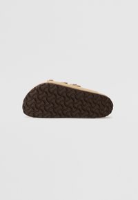 Tan shoe with a textured brown rubber sole featuring wavy patterns. Design includes a low profile with two adjustable straps for fit.