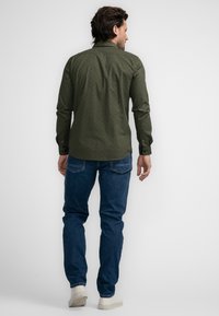 Camisa de patrón verde oscuro con cuello abotonado, combinada con pantalones vaqueros azules y zapatillas blancas, vista desde la parte trasera.