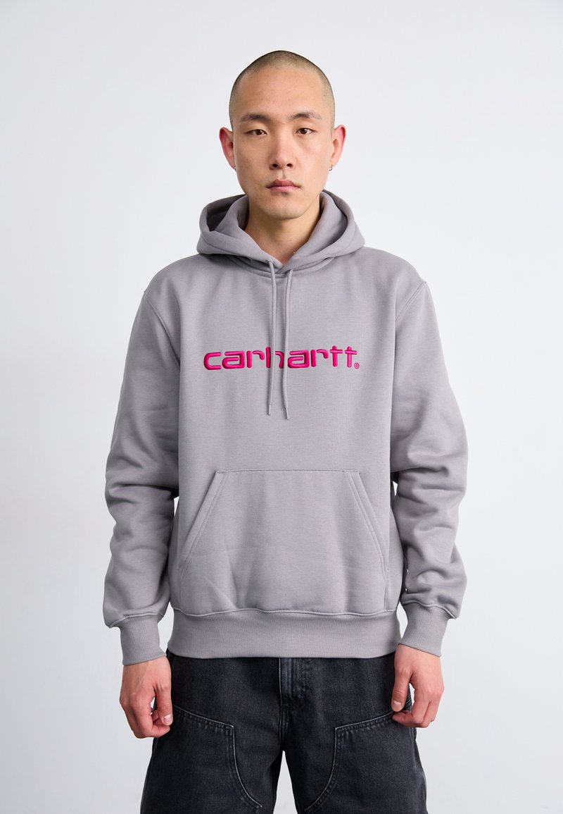 Felpa grigia con il logo 'carhartt.' rosa, cappuccio con lacci, tasca a marsupio, polsini e orlo a costine, e una texture morbida. Indossata da un modello.