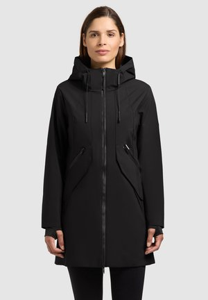 Frau trägt eine lange schwarze Kapuzenjacke mit durchgehendem Reißverschluss und Reißverschlusstaschen, steht vor einem schlichten hellgrauen Hintergrund.