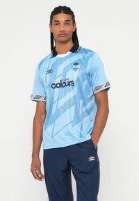Jaunas vyras su ilgomis kasomis, vilkintis mėlyną raštuotą futbolo marškinėlį ir tamsiai mėlynas sportines kelnes su "Umbro" logotipais, stovi prieš baltą foną.