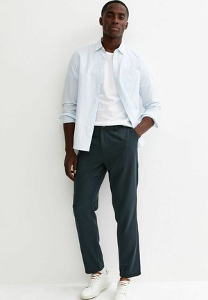 Jonge man staat tegen een witte muur, gekleed in een lichtblauw overhemd over een wit T-shirt, donkere broek en witte sneakers.