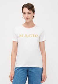 Hvit t-skjorte med gullglittertekst som staver "MAGIC." Korte ermer og klassisk rund hals-design. Brukt med lyseblå jeans.