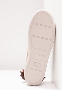 Semelle d'un sneaker beige clair avec un motif de grip texturé, présentée debout contre une surface blanche dans un coin.