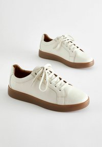 Sneakers bianchi in pelle con una finitura testurizzata, suola in gomma marrone, lacci e dettagli di design minimalista. Accentuati da cuciture chiare.