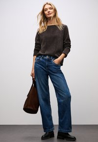 Maglione grigio scuro a trama con scollatura ampia, abbinato a jeans in denim blu larghi. Il modello tiene in mano una borsa in suede marrone e indossa stivali neri.
