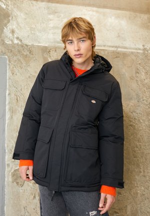 Chaqueta acolchada negra con capucha, cuatro bolsillos frontales y un logo bordado. Presenta un acabado texturizado y un cierre con cremallera.