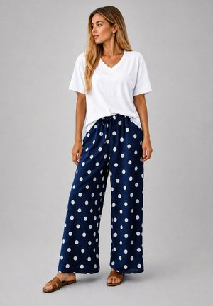 Femme debout portant un T-shirt blanc à col en V, un pantalon large bleu marine à pois blancs et des sandales plates marron, regardant vers sa droite.