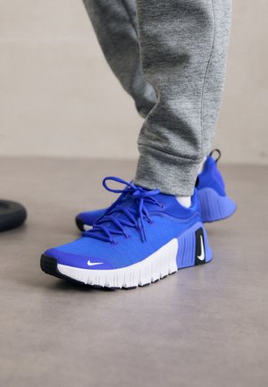 Personne portant des chaussures de course Nike bleues et un pantalon de survêtement gris, debout sur un sol gris lisse avec des détails visibles de la semelle des chaussures.
