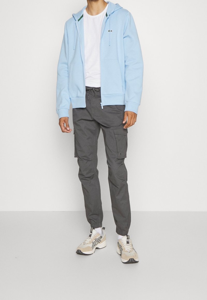 Homme portant un sweat à capuche bleu clair zippé, un t-shirt blanc, un pantalon cargo gris foncé et des baskets blanches beiges, debout devant un fond blanc uni.