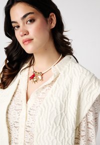 Romige gebreide cardigan over een kant blouse, met een speelse ketting met gouden en rode accenten. Zachte stoffen en gelaagd ontwerp.